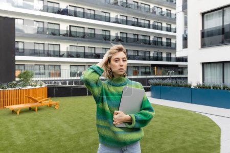 Photo for Blonde woman in green sweater holding laptop and adjusting wavy hair near hotel building in Barcelona - Royalty Free Image