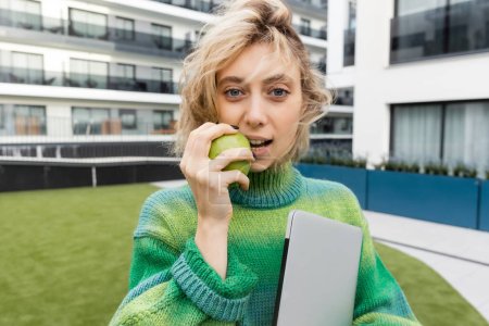 Blondine im Pullover hält Laptop in der Hand und isst grünen Apfel in der Nähe eines Hotelgebäudes in Barcelona 