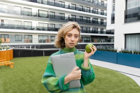 junge Frau im Pullover mit Laptop und grünem Apfel in der Nähe eines Hotelgebäudes in Barcelona 