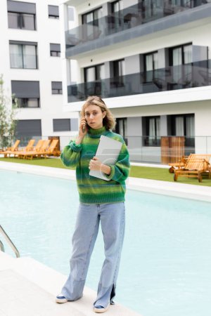 mujer rubia en suéter de pie con portátil mientras habla en el teléfono inteligente cerca de la piscina al aire libre en Barcelona 
