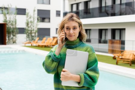 happy woman in sweater standing with laptop while talking on smartphone near outdoor swimming pool 
