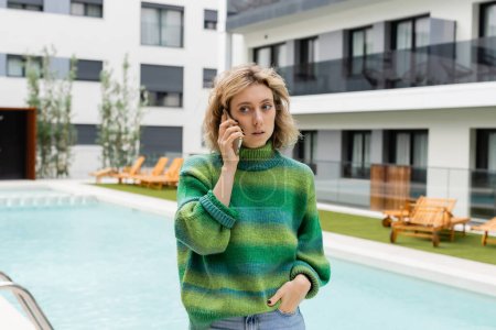 young woman in sweater standing with hand in pocket while talking on smartphone near outdoor swimming pool 