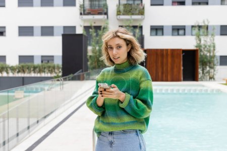 young woman in sweater texting on smartphone near outdoor swimming pool of hotel in Barcelona