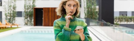 pensive young woman in sweater holding smartphone near outdoor swimming pool of hotel in Barcelona, banner 