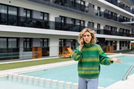 displeased woman in sweater talking on smartphone near outdoor swimming pool 