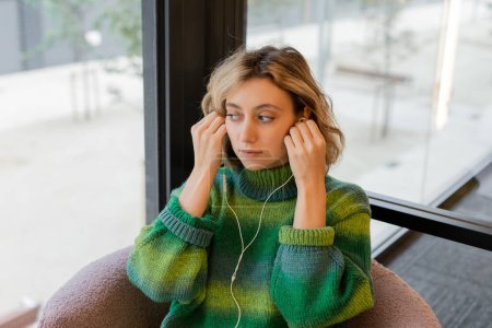 blonde and young woman in green sweater wearing wired earphones while sitting in lobby of hotel in Barcelona 