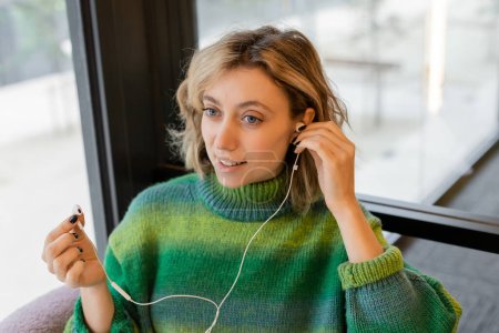Lächelnde junge Frau in grünem Pullover mit kabelgebundenen Kopfhörern, während sie in der Lobby eines Hotels in Barcelona sitzt 