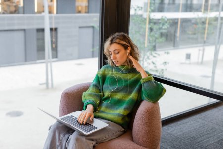 Téléchargez les photos : Jeune femme souriante en pull et écouteurs filaires à l'aide d'un ordinateur portable tout en étant assis dans le hall de l'hôtel à Barcelone - en image libre de droit