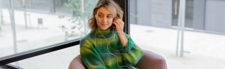 fröhliche Frau in grünem Pullover mit Kopfhörern, während sie in der Lobby eines Hotels in Barcelona sitzt, Banner 