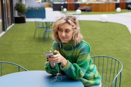 happy young woman in green sweater listening music in wired earphones and holding smartphone 