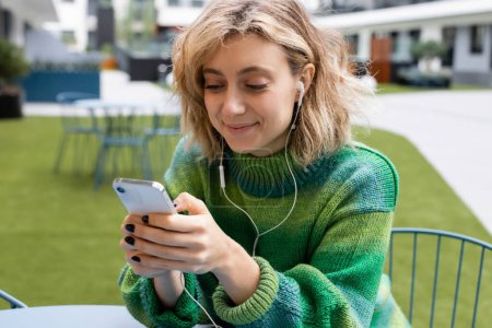 pleased young woman in green sweater listening music in wired earphones and using smartphone 