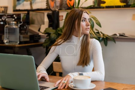 junge Frau sitzt neben Tasse mit Cappuccino und Laptop auf Tisch in Café in Wien 
