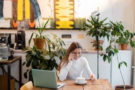 Téléchargez les photos : Blogueur joyeux parler sur smartphone près d'un ordinateur portable à côté de la tasse de cappuccino dans le café - en image libre de droit