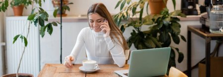 Téléchargez les photos : Femme gaie parlant sur smartphone près d'un ordinateur portable à côté de la tasse de cappuccino dans un café à Vienne, bannière - en image libre de droit