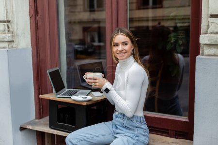 cheerful freelancer holding cup of coffee near laptop with blank screen in cafe while sitting outdoors in Vienna 