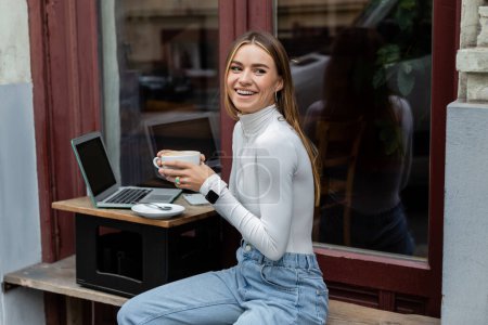 joyful freelancer holding cup of coffee near laptop with blank screen in cafe while sitting outdoors in Vienna 