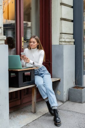 Téléchargez les photos : Femme gaie en utilisant smartphone près d'un ordinateur portable et une tasse de café tout en étant assis dans un café à Vienne - en image libre de droit