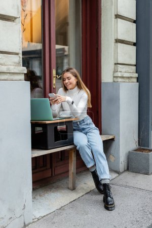 cheerful woman using smartphone near laptop and cup of coffee while sitting in cafe on street in Vienna 