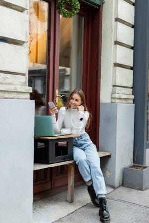 overjoyed woman holding smartphone near laptop and cup of coffee while sitting in cafe on street in Vienna 