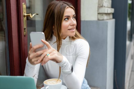 Photo pour Une jeune femme souriante tenant un smartphone près d'une tasse de cappuccino alors qu'elle était assise dans un café à Vienne - image libre de droit