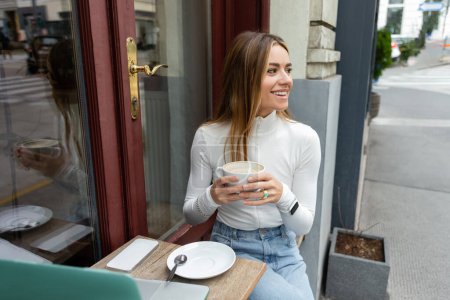 carefree woman holding cup of cappuccino while sitting near gadgets on table in cafe on street in Vienna 