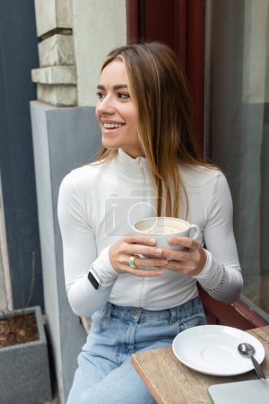carefree woman holding cup of latte while sitting in outdoor cafe on street in Vienna 
