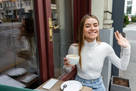 Unbekümmerte Frau hält Cappuccino in der Hand und winkt in Café auf Straße in Wien 