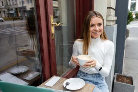 mujer feliz sosteniendo taza de capuchino mientras está sentado en la mesa del bistró con gadgets en la cafetería en la calle en Viena 