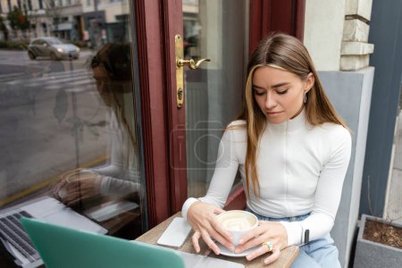 Téléchargez les photos : Jeune femme regardant tasse avec cappuccino tout en étant assis à la table de bistrot près de gadgets dans un café à Vienne - en image libre de droit