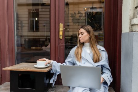 young freelancer in blue trench coat using laptop while reaching cup of coffee in outdoor cafe in Vienna  