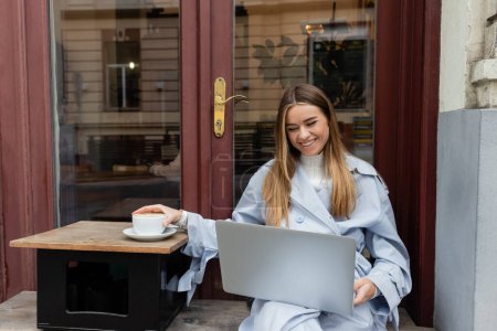 happy woman in blue trench coat using laptop while reaching cup of coffee in outdoor cafe in Vienna  