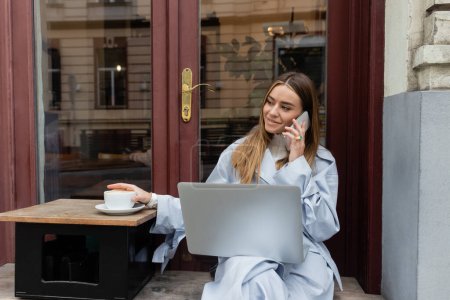 Téléchargez les photos : Femme gaie en trench coat bleu parlant sur smartphone et assis à côté d'une tasse de café dans un café en plein air à Vienne - en image libre de droit