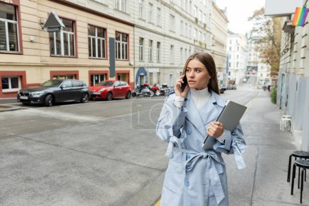 young woman in blue trench coat talking on smartphone while holding laptop on street in Vienna 