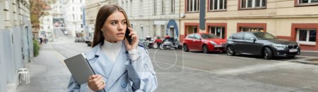 young woman in blue trench coat talking on smartphone while holding laptop on street in Vienna, banner 