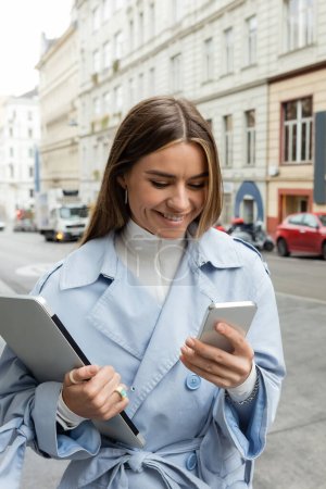 happy woman in blue trench coat using smartphone while holding laptop on street in Vienna 