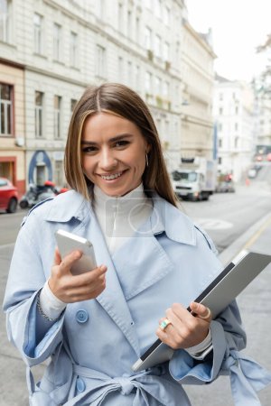 smiling woman in blue trench coat using smartphone while holding laptop on street in Vienna 
