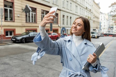 happy woman in blue trench coat taking selfie while holding laptop on street in Vienna 