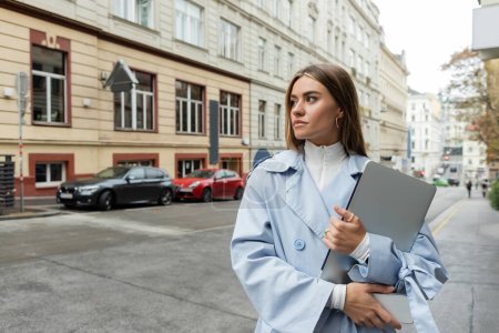 Hübsche Frau in blauem Trenchcoat mit Smartphone und Laptop beim Gassigehen in Wien 