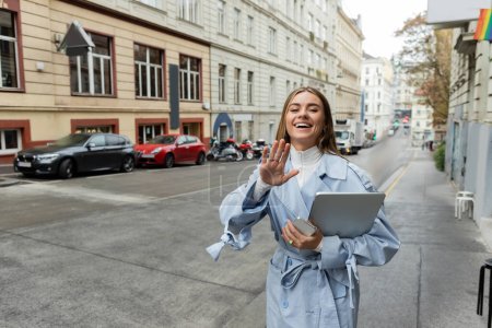 Glückliche Frau im blauen Trenchcoat hält Smartphone und Laptop in der Hand und winkt auf der Straße in Wien 