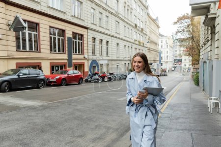 femme souriante en trench coat bleu tenant smartphone et ordinateur portable tout en marchant dans la rue à Vienne 