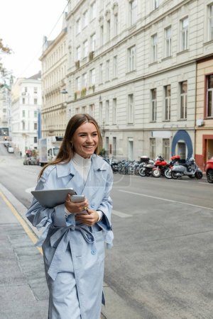 happy woman in trendy trench coat holding smartphone and laptop while walking on street in Austria 