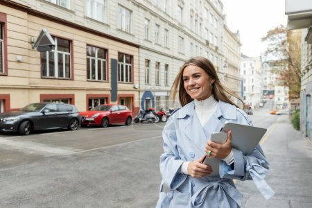 pleased woman in stylish trench coat holding smartphone and laptop while walking on street in Vienna 