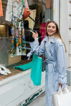 carefree woman in stylish trench coat standing with shopping bags and pointing at showcase of boutique shop in Vienna 