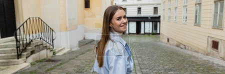 cheerful young woman in blue trench coat walking near buildings on street in Vienna, banner 