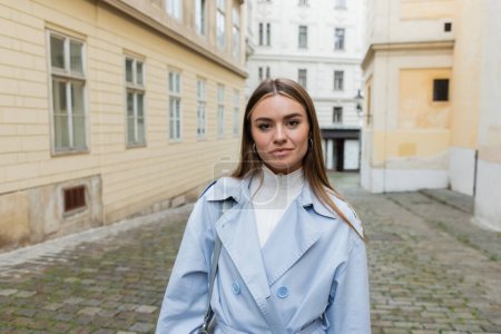 Charmante junge Frau in blauem Trenchcoat steht in der Nähe von Gebäuden auf der Straße in Wien 