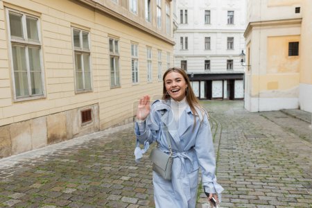 encantadora mujer en gabardina azul saludando de la mano mientras camina con un teléfono inteligente cerca de edificios en Viena 