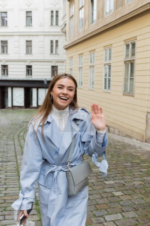 happy woman in blue trench coat waving hand while walking with smartphone near buildings in Vienna 