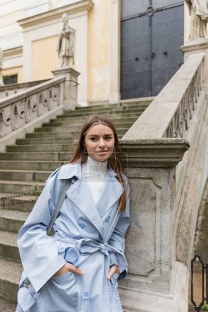 Charmante Frau im blauen Trenchcoat lächelt, während sie mit den Händen in den Taschen in der Nähe der antiken Treppe auf der Straße in Wien steht 