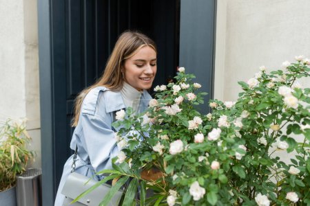 happy young woman in blue trench coat looking at green bush with blooming flowers on street in Vienna 
