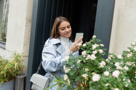 happy young woman in blue trench coat taking photo of green bush with blooming flowers on street in Vienna 